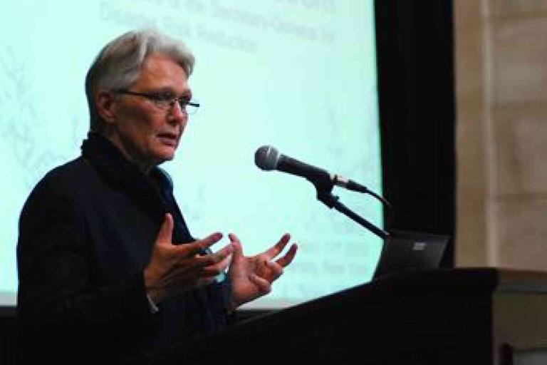 Person speaking at a podium with a microphone, gesturing with hands. A projected presentation screen is visible in the background.
