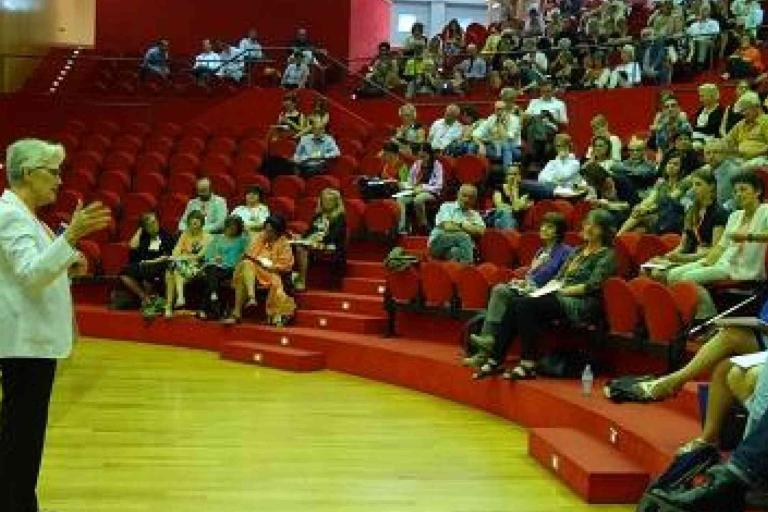 A person stands and speaks to an audience seated in red chairs in a tiered lecture hall.