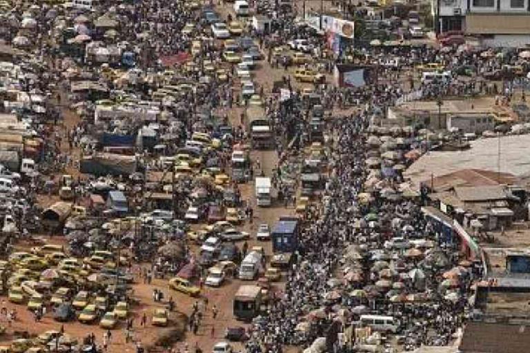 Aerial view of a crowded street filled with vehicles, market stalls, and a large number of people in an urban area.