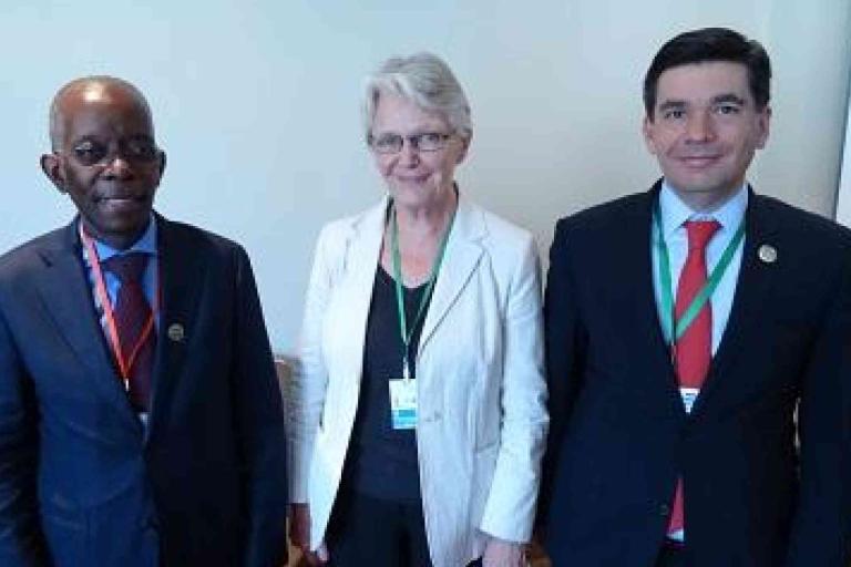 Three people, two men and one woman, stand indoors wearing business attire and conference badges, posing for a group photo.