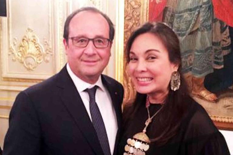 A man in a suit and a woman in a black outfit smiling for a photo in an ornate room with gold accents and a tapestry on the wall.