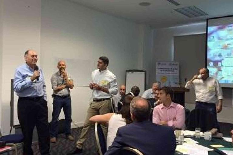 Several people are gathered in a conference room; one man is speaking into a microphone while others listen, with a projector screen and whiteboard in the background.