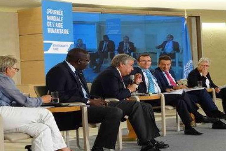 A panel of six people in business attire sit on stage, speaking and listening, with a "Journée Mondiale de l'Aide Humanitaire" banner in the background.