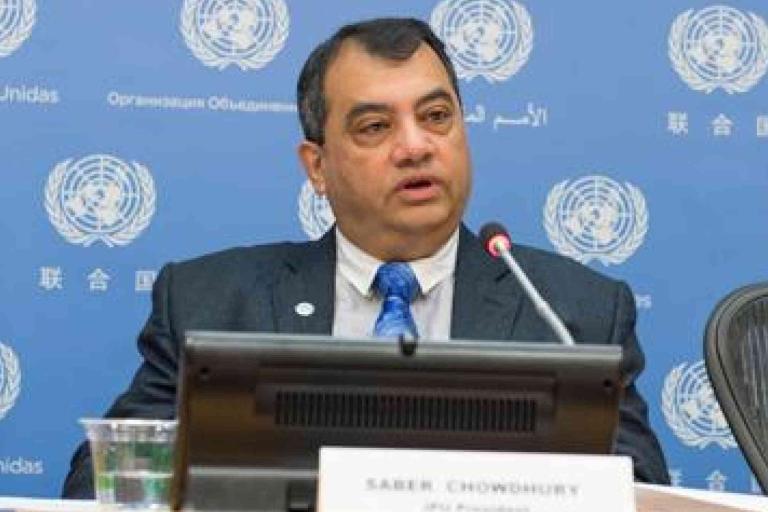 A man in a suit speaks at a United Nations conference table with a nameplate, microphone, and UN logo backdrop.