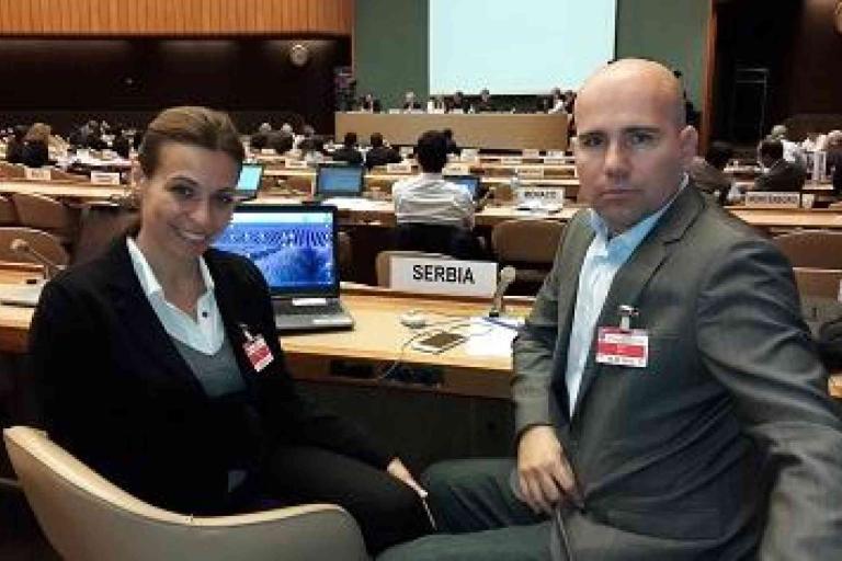 Two people sit at a conference table with "Serbia" nameplate, laptops, and other delegates visible in the background.