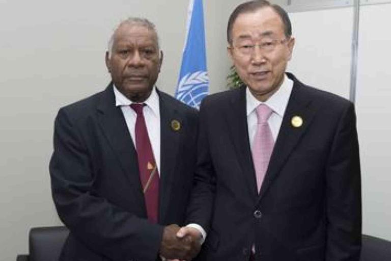 Two men in suits shake hands in front of a United Nations flag in an office setting.