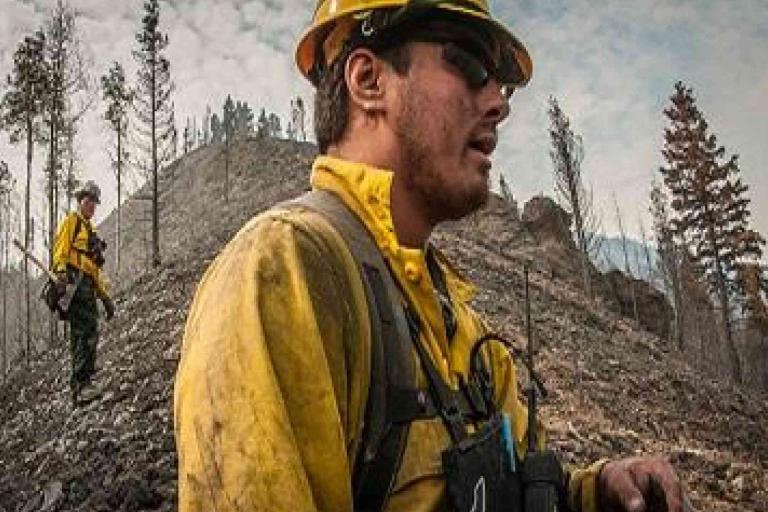 Two firefighters in yellow protective gear stand on rocky terrain with burnt trees in the background, likely assessing wildfire damage.