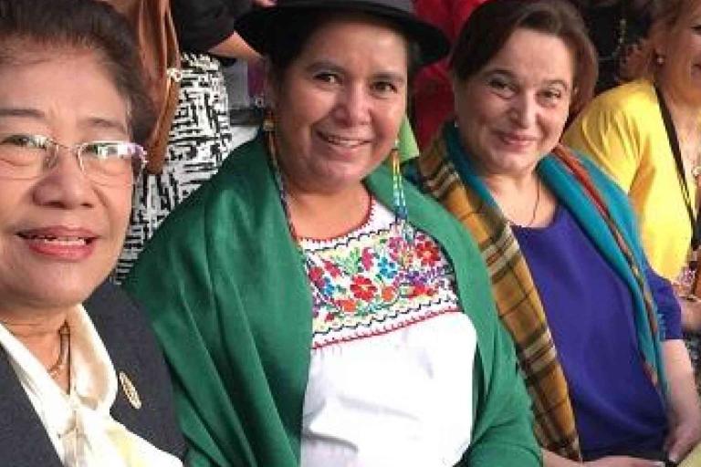 Four women sitting together, smiling at the camera. One woman wears traditional clothing with a green shawl and black hat, while the others are dressed in modern attire.