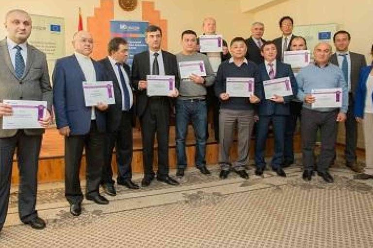 A group of fourteen people stand indoors, several holding certificates, posing for a photo during an award or recognition event.