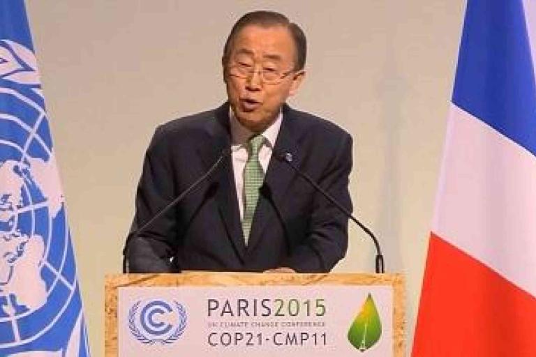 A man in a suit speaks at a podium labeled "Paris 2015 COP21-CMP11" with the UN and French flags in the background.