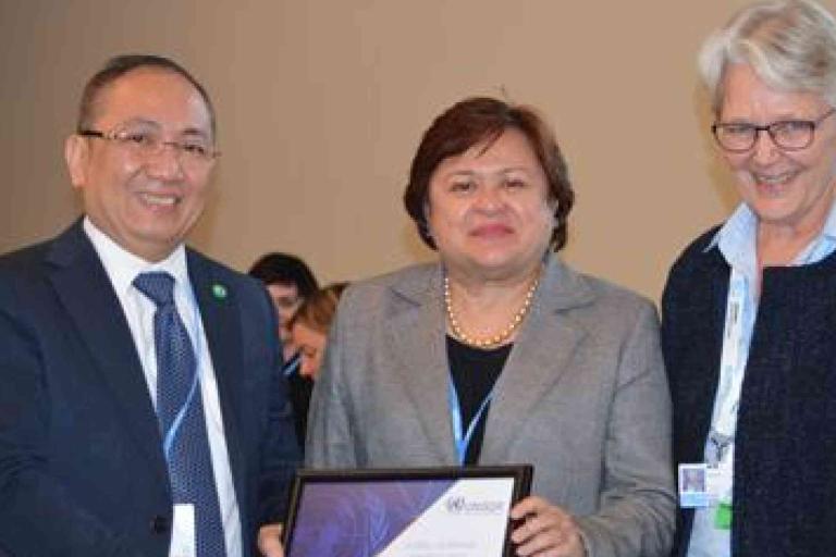 Three adults in business attire stand together indoors, with one person holding a framed certificate and all smiling at the camera.
