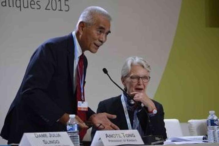A man in a suit speaks at a conference table while a woman with glasses listens beside him. Nameplates and water bottles are on the table.