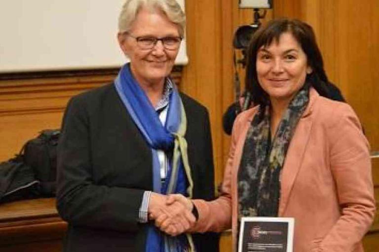 Two women are shaking hands; one is holding a plaque. They are standing in a wood-paneled room with a projector screen in the background.