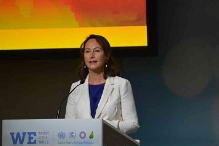 A woman in a white blazer stands at a podium, speaking at an event with a colorful background screen behind her.