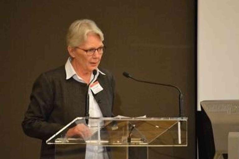An older woman with short gray hair speaks at a podium with a microphone, wearing glasses and a gray blazer, in front of a plain background and projection screen.
