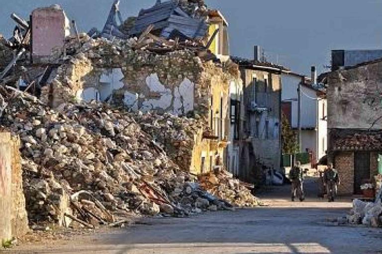 A damaged building with collapsed walls and debris scattered on the street after an apparent earthquake; two people walk in the distance.