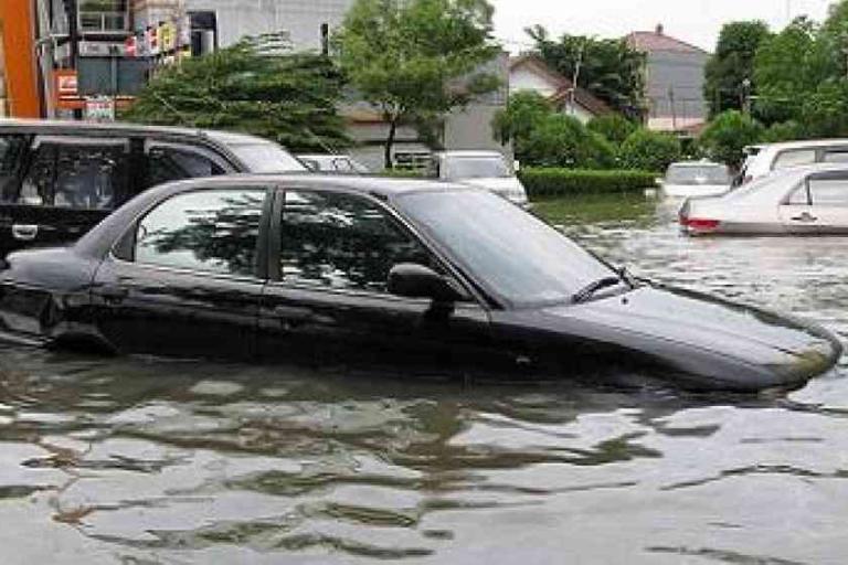 Several cars are partially submerged in floodwater on a city street, with buildings and trees visible in the background.