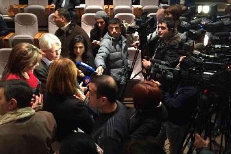 A group of reporters and camera operators surround a person speaking at a press event in an auditorium with empty seats in the background.