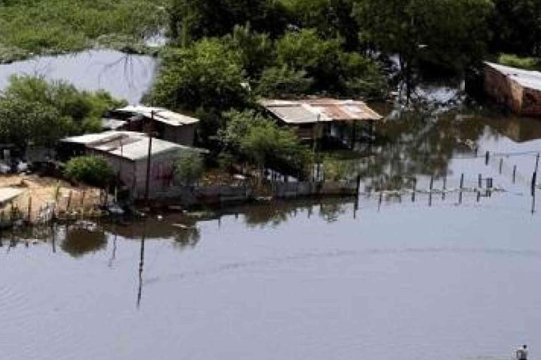 Aerial view of several small houses and trees partially submerged in floodwater, with fences and land barely visible above the waterline.