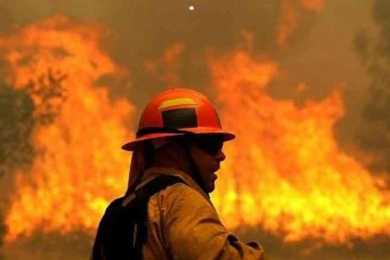A firefighter in safety gear stands in front of a large wildfire, with flames and smoke visible in the background.