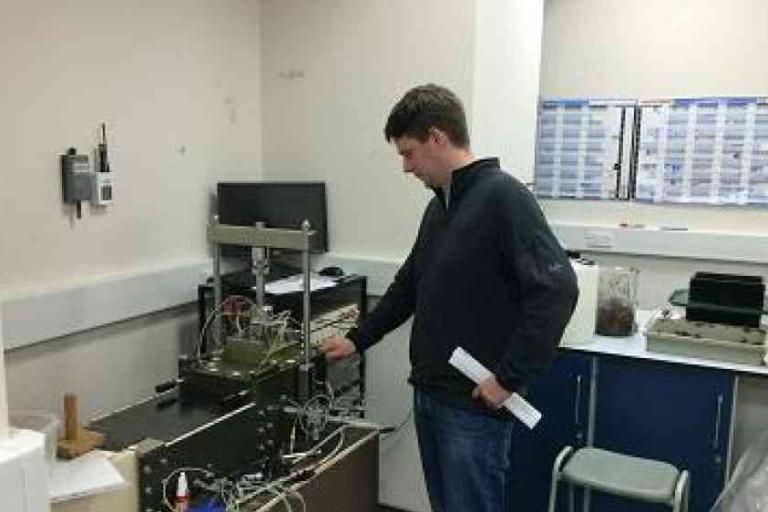 A person stands next to laboratory equipment, adjusting a machine with wires and tubes, in a room with a computer and charts on the wall.