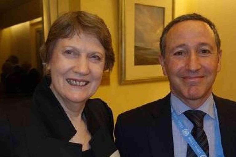 Two people in formal attire stand side by side indoors, smiling at the camera with framed artwork on the wall behind them.