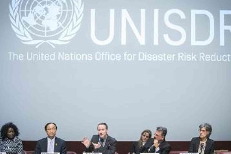 Six panelists sit at a long table in front of a large UNISDR logo and text reading "The United Nations Office for Disaster Risk Reduction.