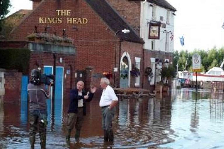 A cameraman films two men talking while standing in floodwater outside The King’s Head pub; water surrounds the building and a boat is visible in the background.