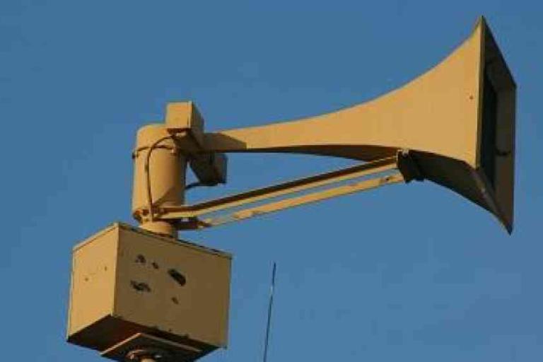 A large outdoor warning siren mounted on a metal pole against a clear blue sky.