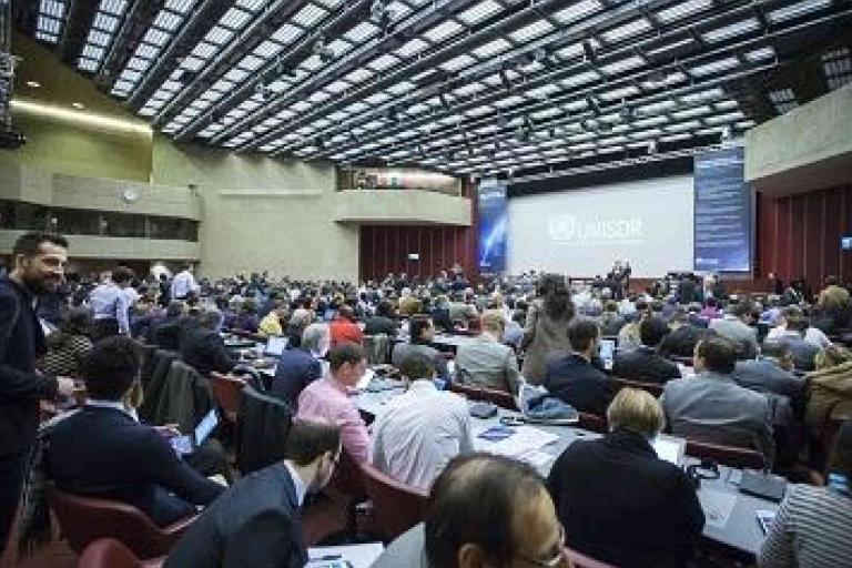 A large conference hall filled with seated attendees facing a stage with a presentation screen displaying the UNISDR logo.