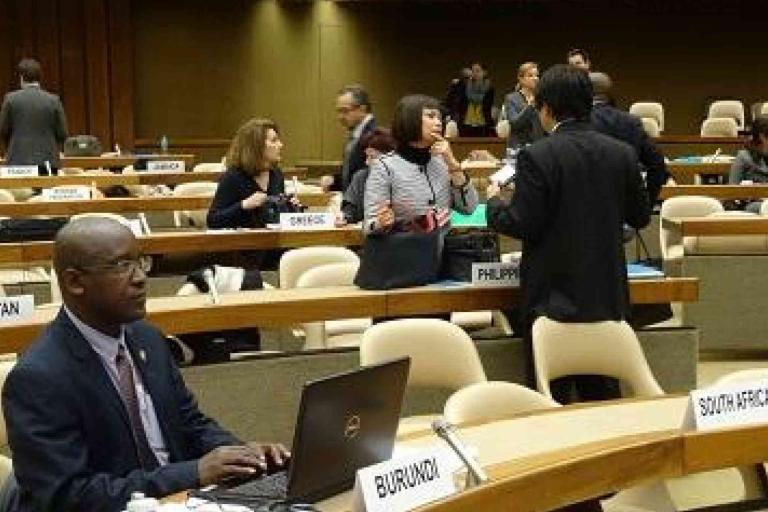 Delegates from various countries sit and converse in a conference room with country nameplates visible, including Burundi, South Africa, and Bhutan.