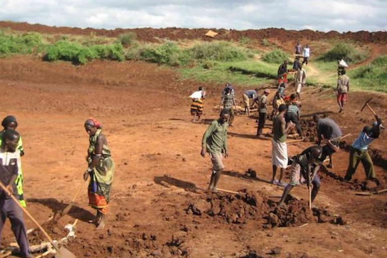 A group of people work with tools on a large, barren, reddish-brown expanse of land under a cloudy sky.