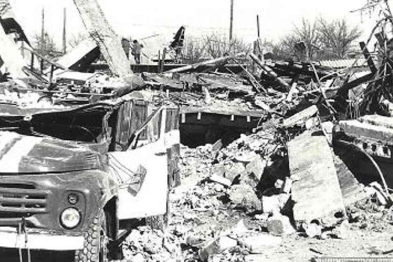 A damaged truck is parked amid rubble and collapsed concrete structures in a heavily destroyed area.