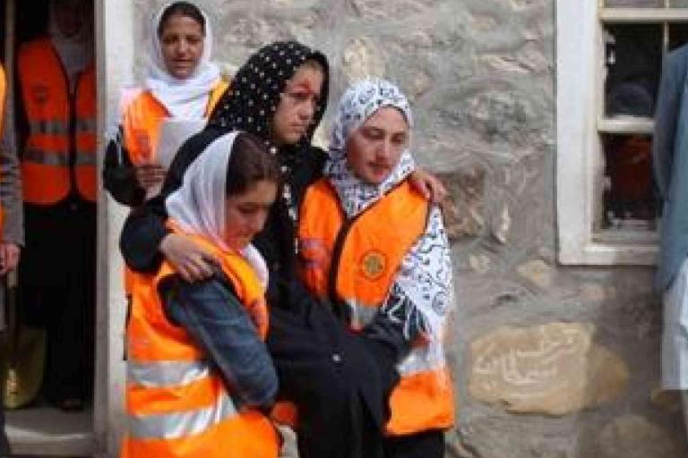 Several women wearing orange safety vests assist and support another woman outside a stone building.