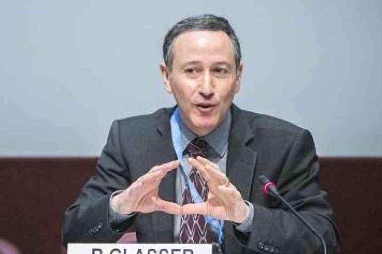 A man in a suit speaks into a microphone at a conference, gesturing with his hands. A nameplate reading "P. Glasser" is visible in front of him.