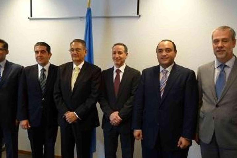 Six men in suits stand side by side in front of a wall with a blue flag, posing for a formal group photo.