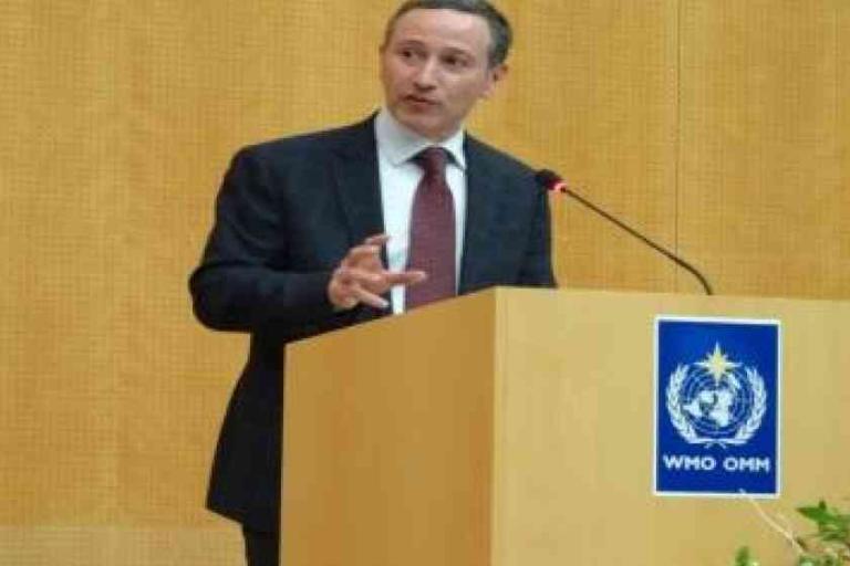 A man in a suit speaks at a podium with a microphone; the podium displays the WHO emblem against a wood-paneled background.