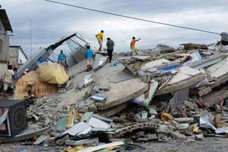 Several people stand and search on top of the rubble of collapsed buildings after an earthquake, with debris scattered around.