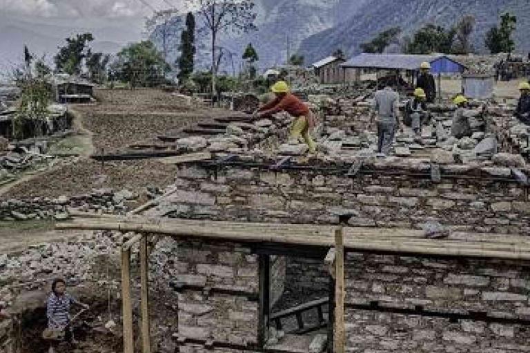 Construction workers wearing helmets build a stone house in a rural, mountainous area, with scattered debris and unfinished structures around them.