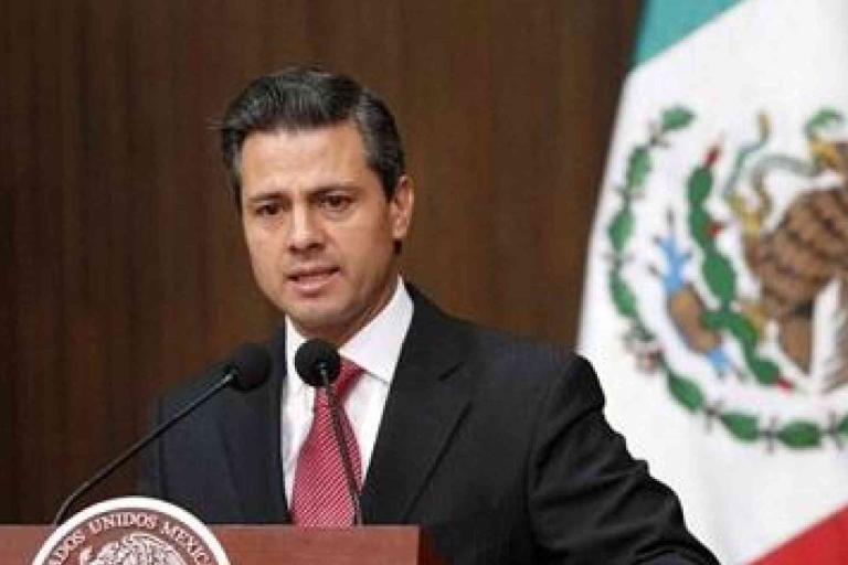 A man in a suit speaks at a podium with microphones, with a Mexican flag visible in the background.