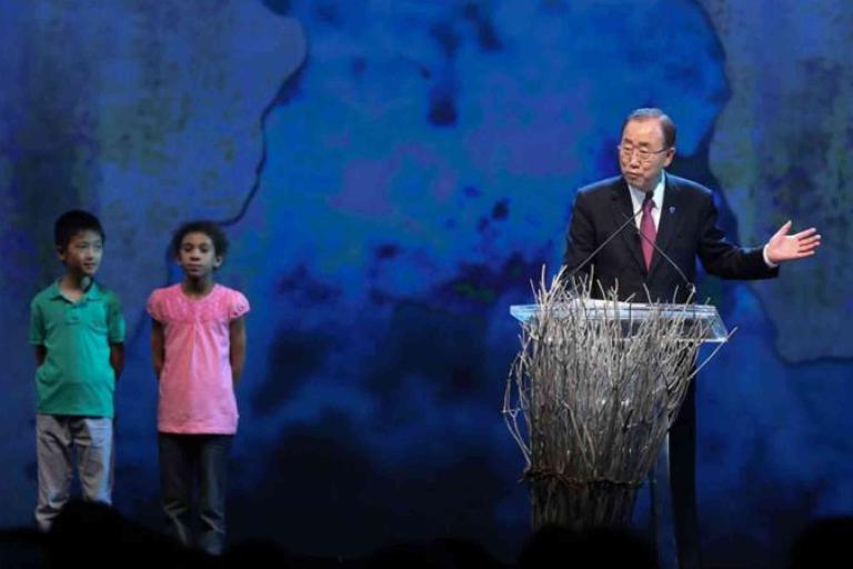 A man speaks at a podium decorated with branches, while a boy and a girl stand on stage beside him against a blue background.