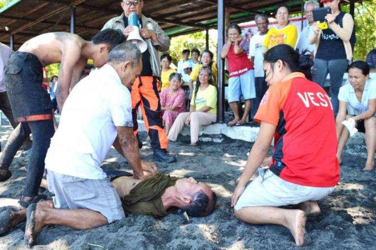 A group of people assists a man lying on the ground at a beach, while bystanders watch in the background.