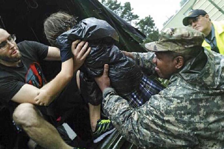 A man and a soldier help a child wearing a plastic cover climb into a vehicle as another person in a yellow raincoat watches.
