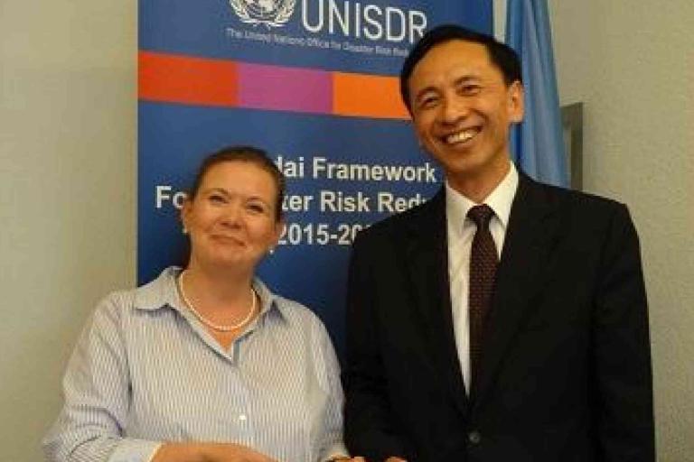 Two people stand in front of a UNISDR banner, smiling and shaking hands at an event related to disaster risk reduction.