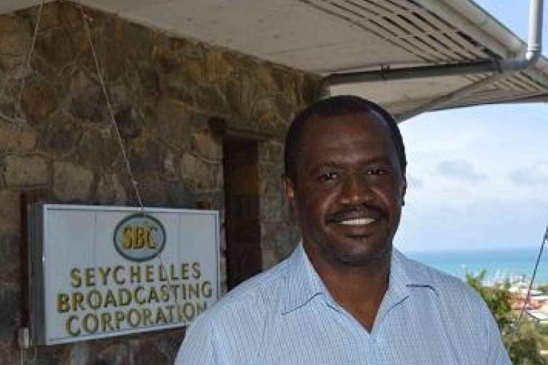 A man stands in front of a building with a sign reading "Seychelles Broadcasting Corporation," with a coastal view in the background.