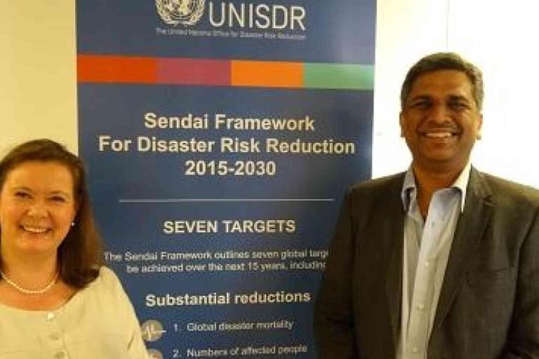 Two people stand and smile in front of a banner for the Sendai Framework for Disaster Risk Reduction 2015-2030 by UNISDR, outlining its seven targets and substantial reductions.