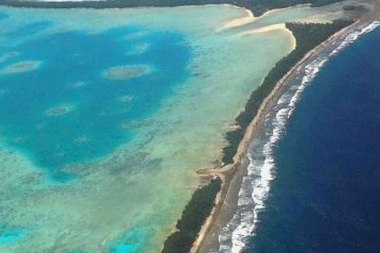 Aerial view of an island with a sandy shoreline, turquoise shallow waters on one side, and deep blue ocean with waves on the other side.
