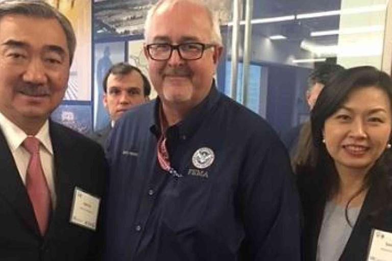 Three people pose for a photo at an indoor event; two wear business attire with name tags, and one wears a FEMA shirt and lanyard.