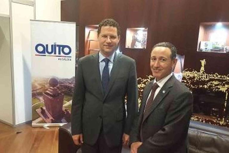 Two men in suits stand indoors near a banner that reads "Quito, Ecuador." Shelves with objects and a cityscape wall design are visible in the background.