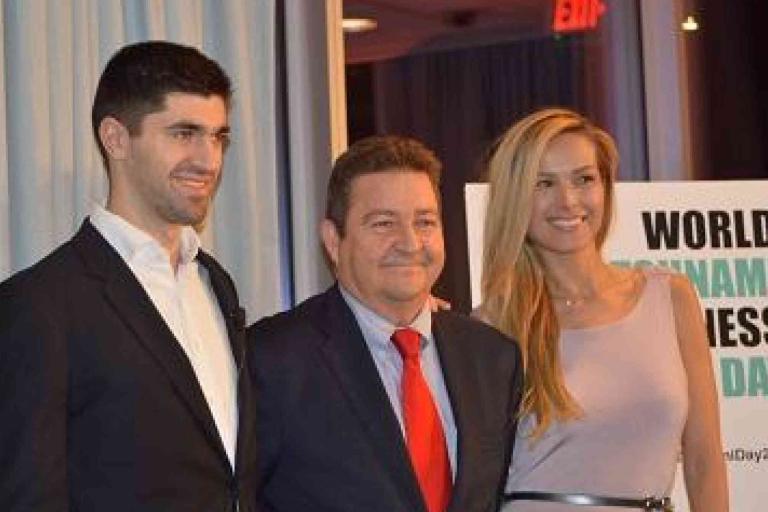 Three people in formal attire stand together, posing for a photo at an event. A sign in the background reads "World Tsunami Awareness Day.